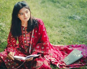 Trends in Women wear in 2026 A young woman in a vibrant traditional outfit reading a book while sitting on grass in Khulna, Bangladesh.